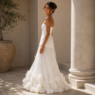 A woman wearing a strapless white wedding dress with a structured bodice and a voluminous skirt made of layered ruffles, standing in a sunlit architectural setting with stone columns and a large potted plant.