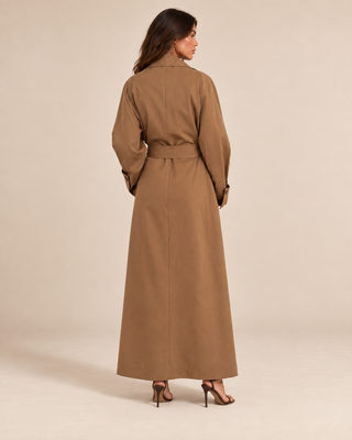 A woman viewed from the back wearing a long tan trench-style dress or maxi coat with a waist tie, standing against a plain warm beige studio background.