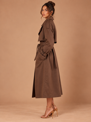 A woman modeling a brown trench coat with a matching waist tie, shown in profile against a solid tan studio backdrop.