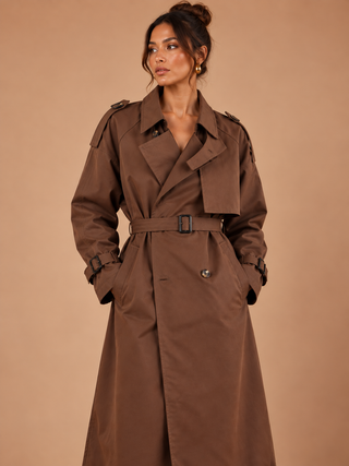 A woman modeling a brown trench coat against a solid warm beige studio background.
