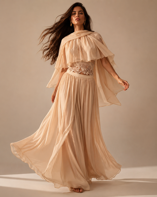 A woman modeling a flowing champagne beige maxi dress with a draped cape-style collar, sheer flutter sleeves, a lace-embellished waist, and a voluminous pleated skirt, set against a solid warm beige studio background.