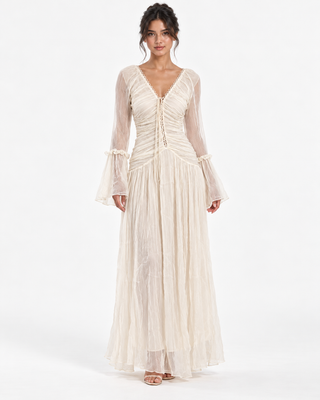 A woman modeling a long-sleeved cream maxi dress with a gathered, ruched bodice, deep V-neckline, and flared bell sleeves against a plain white studio background.