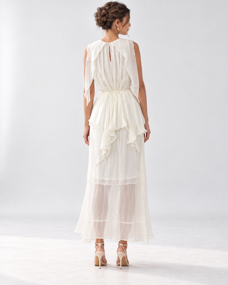 A woman viewed from the back wearing a sleeveless cream maxi dress with a draped, layered ruffle design and a keyhole back, paired with beige high-heeled sandals against a light gray studio background.