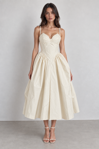 A woman modeling a cream-colored midi dress with spaghetti straps, a sweetheart neckline, a structured corset-style bodice, and a voluminous gathered skirt, paired with white strappy high-heeled sandals against a neutral studio backdrop.