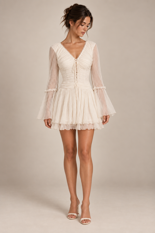 A woman modeling a short cream-colored mini dress with long sheer bell sleeves, a V-neckline, and a gathered corset-style bodice with lace-up front details, set against a solid beige studio background.