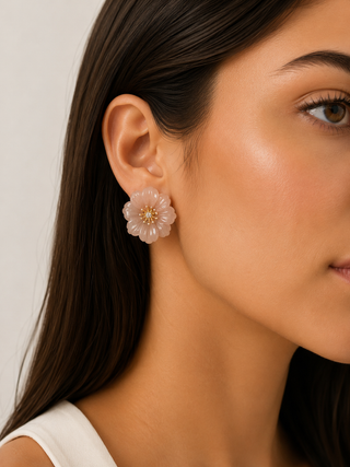 A close-up of a woman's ear wearing a translucent pale pink flower-shaped stud earring with a small gold center, against a light neutral background.