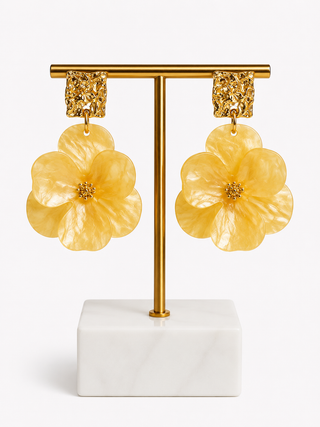 A pair of yellow flower-shaped dangle earrings with textured gold tops, displayed on a gold T-stand with a white marble base against a light gray background.