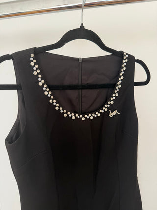 A sleeveless black dress hanging on a black hanger against a plain light background. The dress features a scoop neckline embellished with a row of white pearls and a small gold script logo on the right side.
