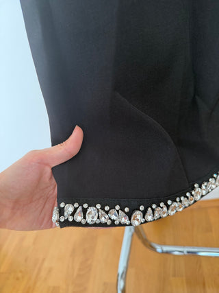 A close-up of a black garment sleeve or hem being held by a hand, showing a decorative border of clear rhinestones and crystals along the edge. A light wood floor and part of a chair are visible in the background.