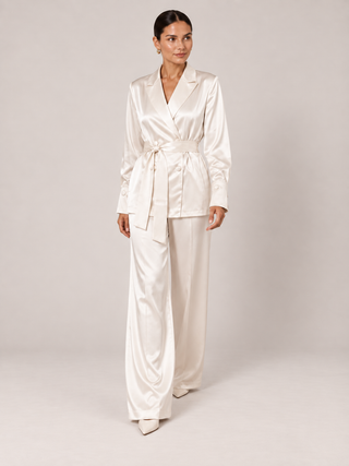 A woman modeling a matching cream satin two-piece set consisting of a belted wrap jacket and wide-leg trousers, photographed against a neutral light gray studio background.