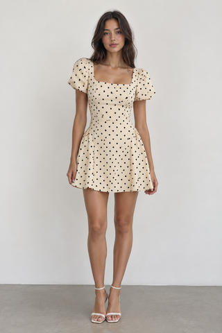 A woman modeling a short cream-colored dress with black polka dots, featuring puff sleeves and a square neckline, standing against a plain light gray wall.