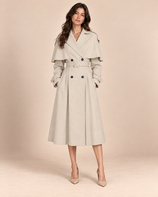 A woman models a light beige double-breasted trench coat with a matching belt, styled with beige high heels against a solid beige studio backdrop.
