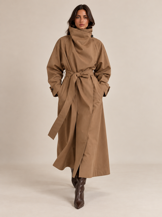 A model wearing a long, high-neck camel brown trench coat with a self-tie belt and dark brown leather ankle boots, standing against a solid light beige studio background.