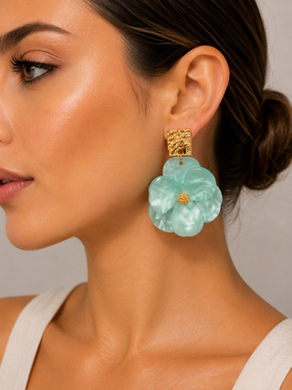 A close-up of a woman wearing a large, light aqua blue flower-shaped drop earring with a textured gold top and a small gold center.