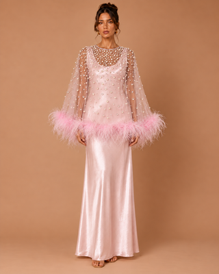 A woman models a floor-length light pink gown with a sheer beaded cape overlay and a pink feather trim at the sleeves, set against a solid warm tan studio background.