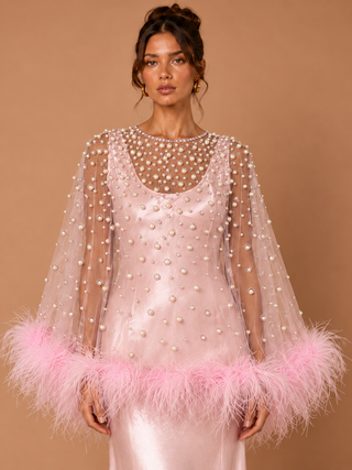 A model wears a light pink satin dress with a sheer embellished cape featuring pearl-like beads and a fluffy pink feather trim at the hem, set against a solid warm tan studio background.
