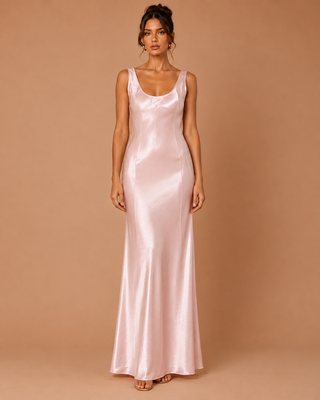 A woman modeling a sleeveless, floor-length satin slip dress in a soft blush pink color, standing against a solid warm tan studio backdrop.