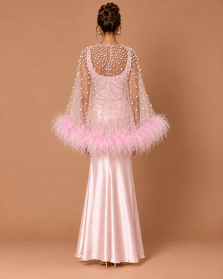 A woman stands with her back to the camera wearing a formal light pink gown with a sheer beaded cape and a fluffy pink feather trim at the sleeves, set against a solid tan studio backdrop.