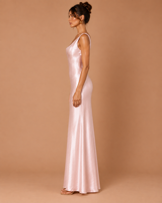 A woman stands in profile wearing a floor-length satin slip gown in a soft blush pink color, set against a solid warm tan studio backdrop.