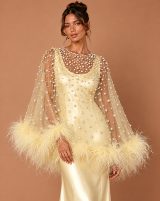 A model wears a champagne-colored evening gown with sheer beaded cape sleeves and fluffy feather trim at the cuffs, set against a solid warm brown studio background.