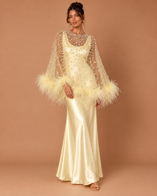 A woman models a floor-length cream satin gown with a sheer embellished cape overlay and feathered cuffs, set against a solid tan studio backdrop.