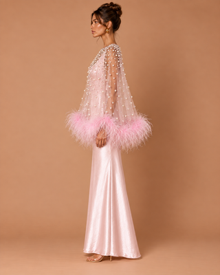A woman models a floor-length light pink satin gown with a sheer embellished overlay and fluffy pink feather trim at the sleeves, posed against a solid warm tan studio backdrop.