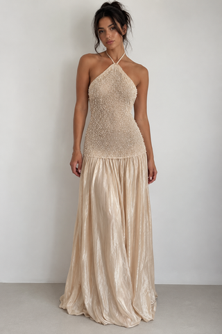 A woman modeling a champagne-colored halter-neck maxi dress with a beaded bodice and a flowing satin skirt, standing against a plain light gray wall.