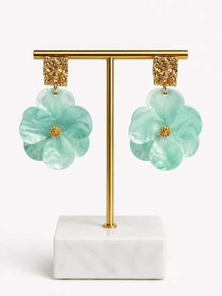 A pair of dangling flower earrings displayed on a gold T-stand with a white marble base against a clean white background. The earrings feature layered mint green flower petals with textured gold square studs.
