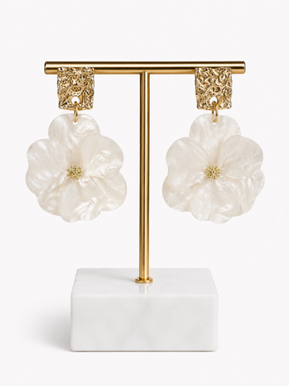 A pair of white flower-shaped dangle earrings with textured gold ear clips, displayed on a gold T-stand with a white marble base against a clean white background.
