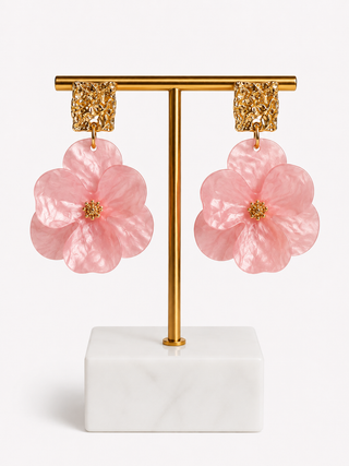 A pair of dangling flower earrings displayed on a gold T-stand with a white marble base against a clean white background. The earrings feature large, layered pink flower petals with textured gold centers and rectangular gold hooks.
