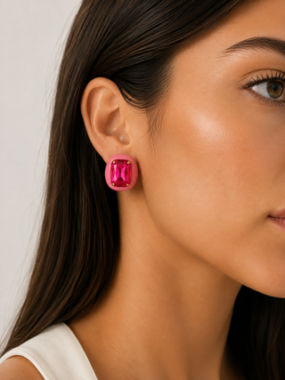 A close-up of a woman's ear wearing a bright pink rectangular stud earring with a faceted gemstone, set against a clean light gray background.
