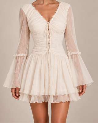 A woman modeling a short cream-colored mini dress with long sheer bell sleeves, a gathered corset-style bodice, lace-up front, and a ruffled tiered skirt against a plain beige studio background.