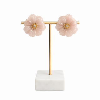 A pair of light pink flower-shaped stud earrings with gold centers, displayed on a gold T-stand with a white marble base against a white background.