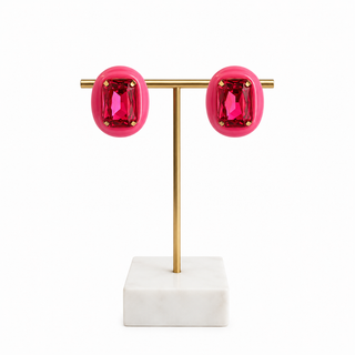 A pair of bright pink rectangular stud earrings with gold-toned settings, displayed on a gold T-stand with a white marble base against a white background.