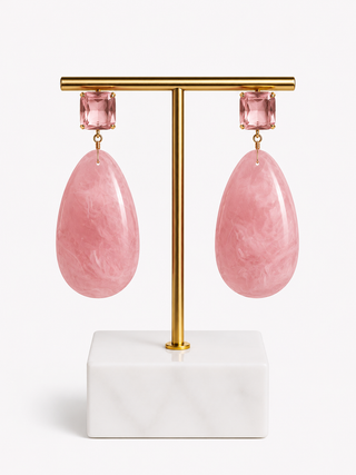 A pair of pink teardrop-shaped drop earrings with rectangular pink stones at the top, displayed on a gold T-bar stand with a white marble base against a light gray background.