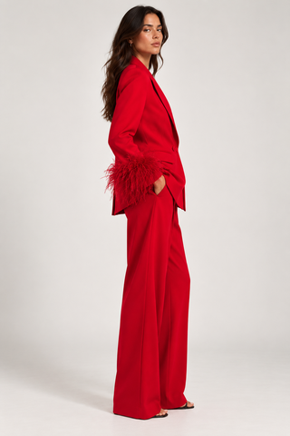 A woman modeling a bright red two-piece suit with wide-leg trousers and a blazer featuring dramatic feathered cuffs, photographed against a plain light gray studio background.