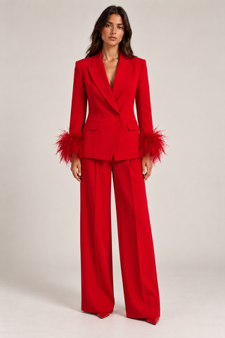 A model stands against a light beige studio background wearing a bright red tailored suit with wide-leg trousers and long sleeves featuring dramatic feathered cuffs.