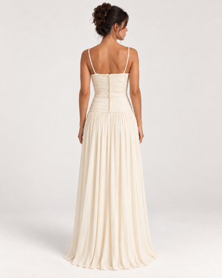 A woman shown from the back wearing a floor-length cream formal dress with thin straps, a fitted ruched bodice, and a flowing pleated skirt against a light gray studio background.