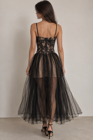 A woman shown from the back wearing a black lace and tulle formal dress with thin straps, a fitted lace bodice, and a voluminous sheer tulle skirt, photographed against a plain light beige studio wall.