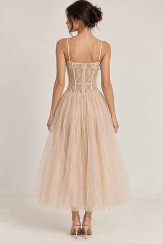 A woman shown from the back wearing a sleeveless tulle dress with thin straps, a lace corset-style bodice, and a full flowing skirt in a light blush nude color, photographed against a plain light studio background.