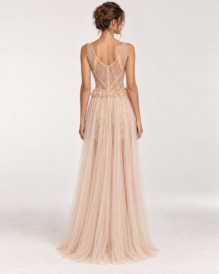 A woman viewed from the back wearing a floor-length formal gown in a pale blush pink tone. The dress has a sheer lace open-back bodice, thin straps, a floral waistband, and a flowing tulle skirt on a plain light studio background.