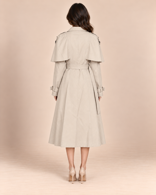 A woman viewed from the back wearing a light beige trench coat with a belted waist and shoulder epaulets, paired with nude high heels, against a solid pale peach studio background.