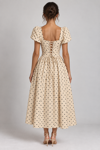 A woman viewed from the back wearing a cream midi dress with small black polka dots, puff sleeves, a square neckline, and a lace-up open back, paired with nude high heels against a light gray studio background.
