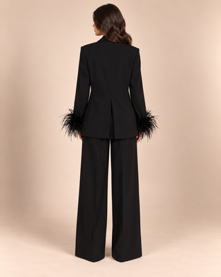 A model viewed from the back wearing a black tailored pantsuit with wide-leg trousers and a blazer featuring dramatic feathered cuffs, set against a solid beige studio background.