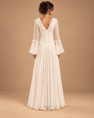 A woman viewed from the back wearing a floor-length ivory wedding dress with long sheer bell sleeves, a deep V back, gathered ruching at the waist, and a flowing pleated skirt, standing against a warm tan studio backdrop.