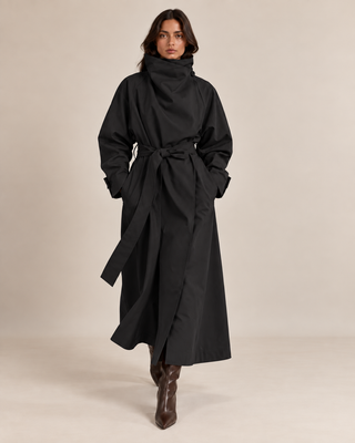 A model wearing a long black wrap-style coat with a high folded collar and self-tie belt, paired with brown pointed-toe ankle boots, against a plain beige studio background.