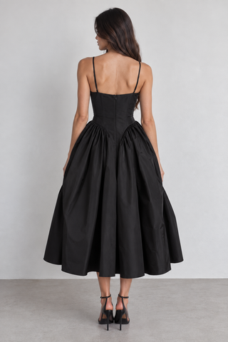 A woman viewed from behind wearing a black midi dress with thin spaghetti straps, a structured corset-style waist, and a full gathered skirt, paired with black high heels against a light gray studio background.