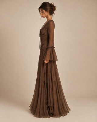 A woman modeling a floor-length brown sheer mesh gown with long flared sleeves and a gathered waist, photographed against a plain warm beige studio background.
