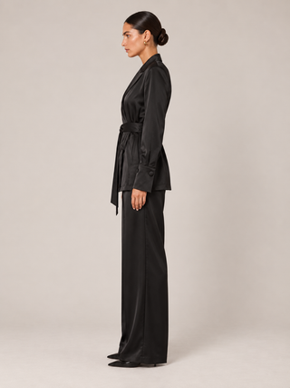 A woman stands in profile wearing a black satin belted wrap jacket and matching wide-leg trousers against a plain light beige studio background.