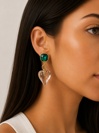 A close-up of a woman's ear wearing a gold drop earring with a green square gemstone and a dangling clear heart-shaped crystal.